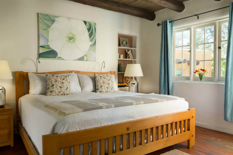 Bed with white bedding and pine bed in bright room with plastered walls, beams in ceiling, Lamps on stands on both sides.