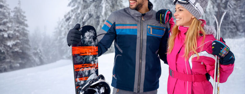 Cute yong couple in ski gear holding a snowboard and skis surrounded by snow in Northern New Mexico