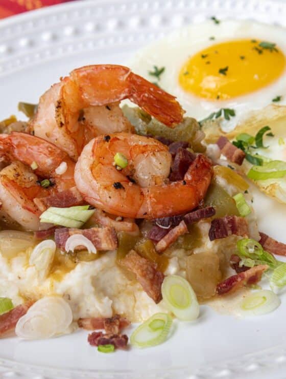 Plate of grits topped with green chile shrimp sauce, shrimp, bacon bits, green onion, and a side of eggs.