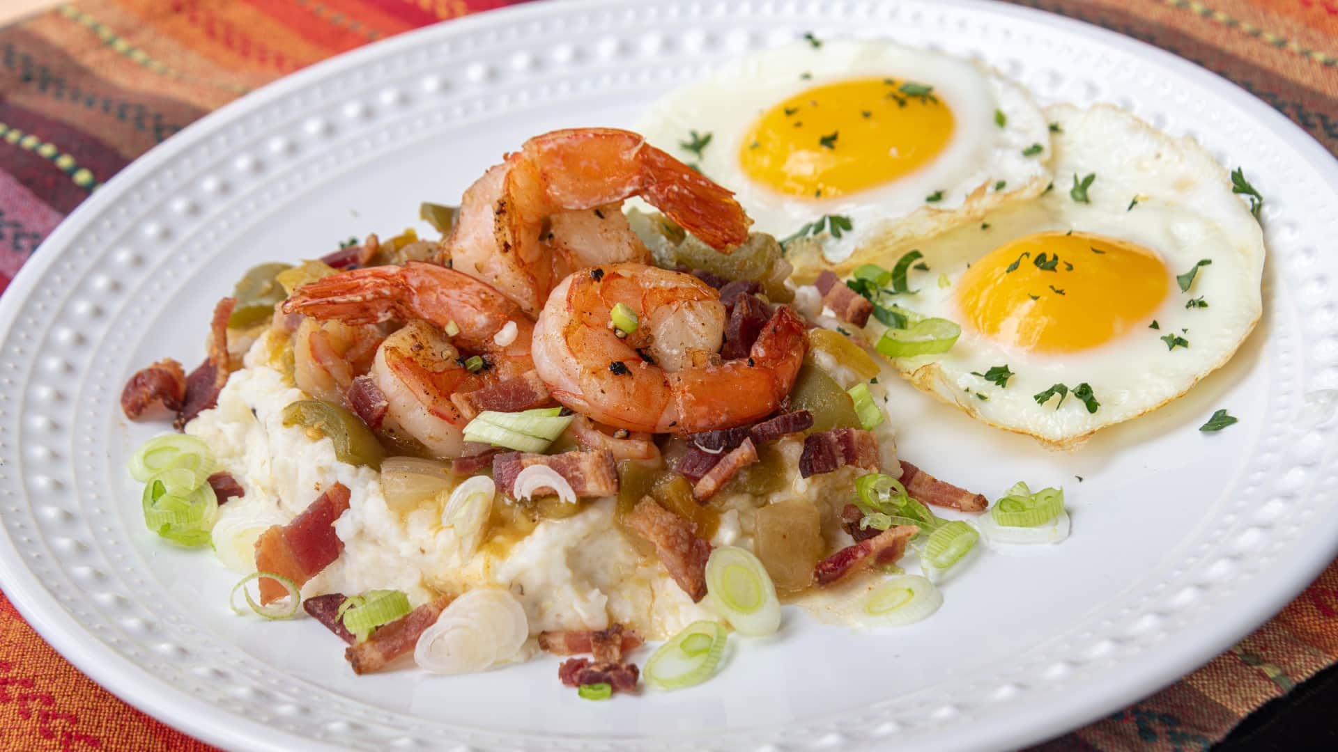 Plate of grits topped with green chile shrimp sauce, shrimp, bacon bits, green onion, and a side of eggs.