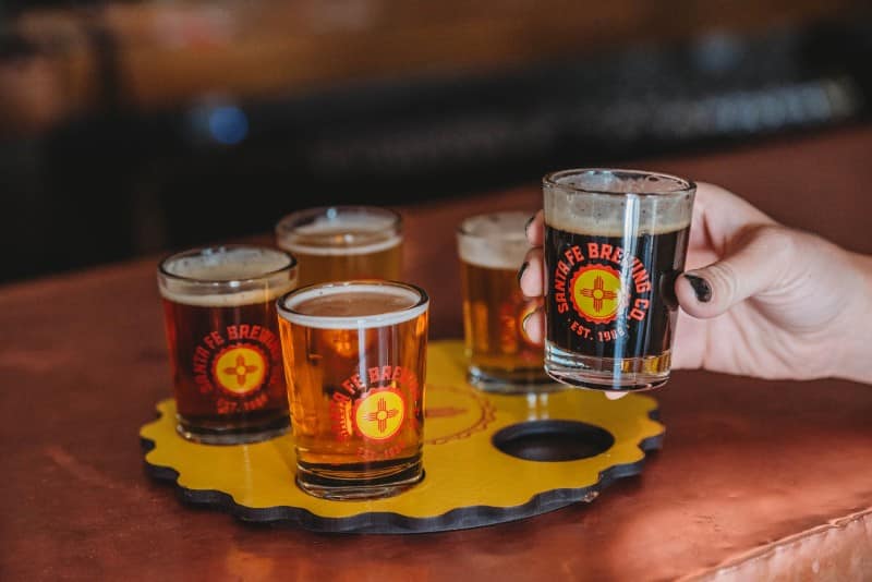 Santa Fe Brewing Co. sampler close up|two men enjoying a drinks and food|single malt whisky with appetizers|girls night out drinking wine|drinking and cheers in group setting|couple taking selfie with beers|comfortable rooms at the Inn|owner of Santa Fe Spirits pouring a drink|up close shot of drink at Dragon Room Lounge