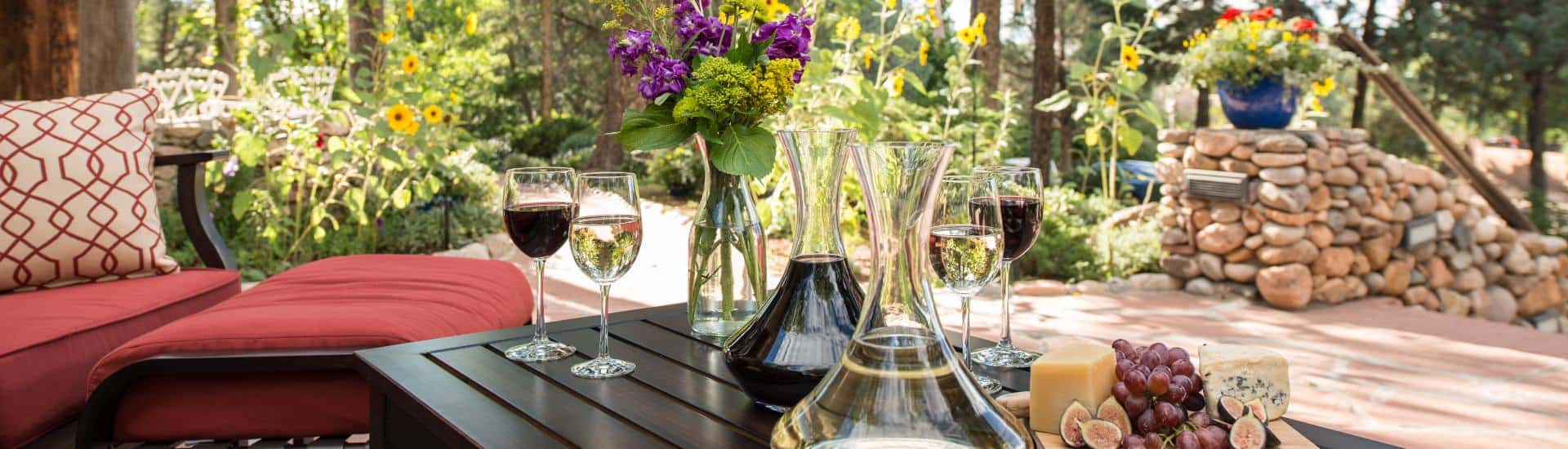 Wine decanters and glasses on wooden patio table outdoors in dappled light from garden.