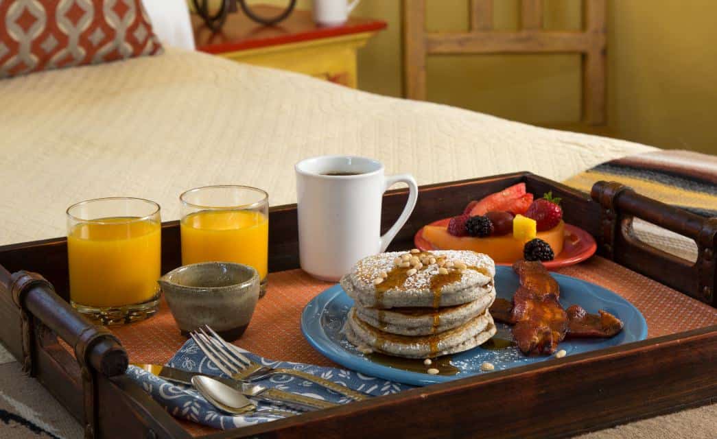 Breakfast tray with pancakes, bacon, fruit, juice and coffee on a bed with a cream coverlet.