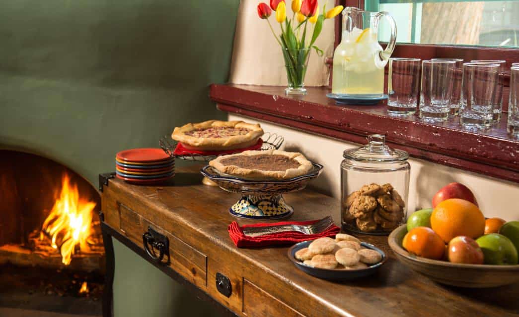 Pies, cookies, and fruit set out on a bar in a room with a green adobe fireplace.