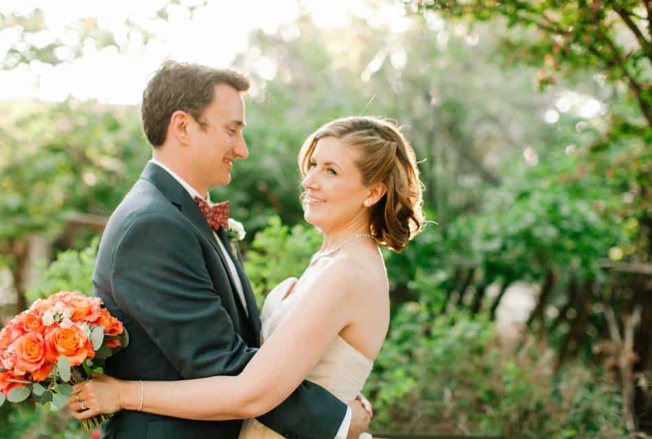 Man in a suit embracing a woman in a wedding dress holding an orange rose bouquet in a garden.