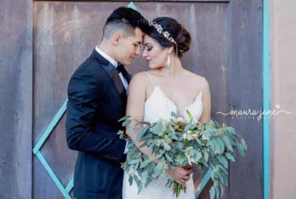 Brunette man in black tuxedo embraces brunette woman in white wedding dress with bouquet of greenery.