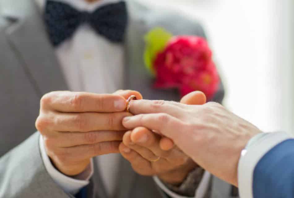 Groom in grey tuxedo placing a wedding band on hand of groom in blue tuxedo.