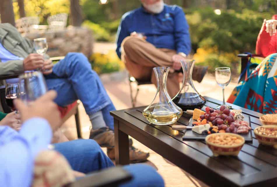 Group of people holding wine glasses relaxing outdoors around a table with wine and snacks.