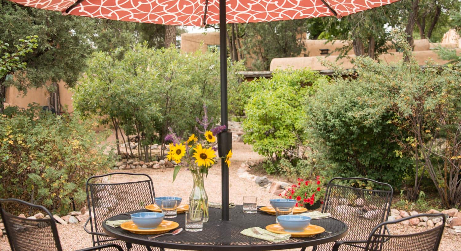 Wrought iron patio table and chairs, blue bowls and orange plates, yellow and purple flowers in a glass vase, and green bushes in the background