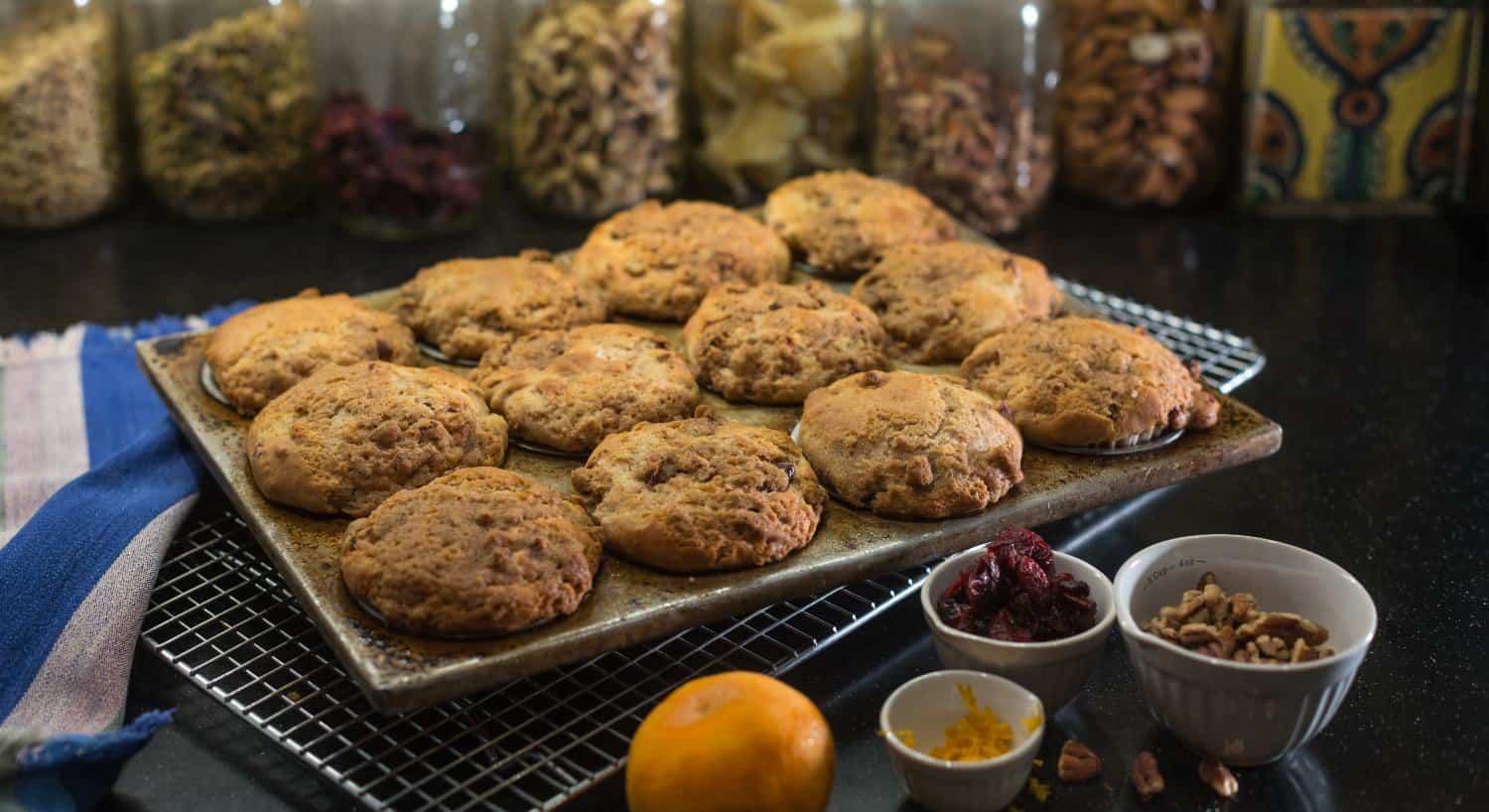 Close up view of baked muffins in the tin with small bowls filled with nuts, dried fruit, and orange zest