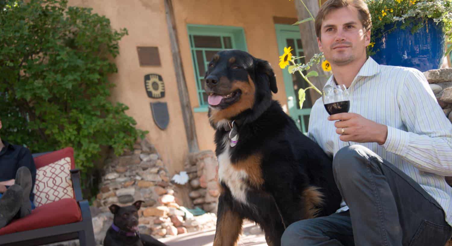 Man with striped shirt holding glass of red wine sitting by black and brown dog