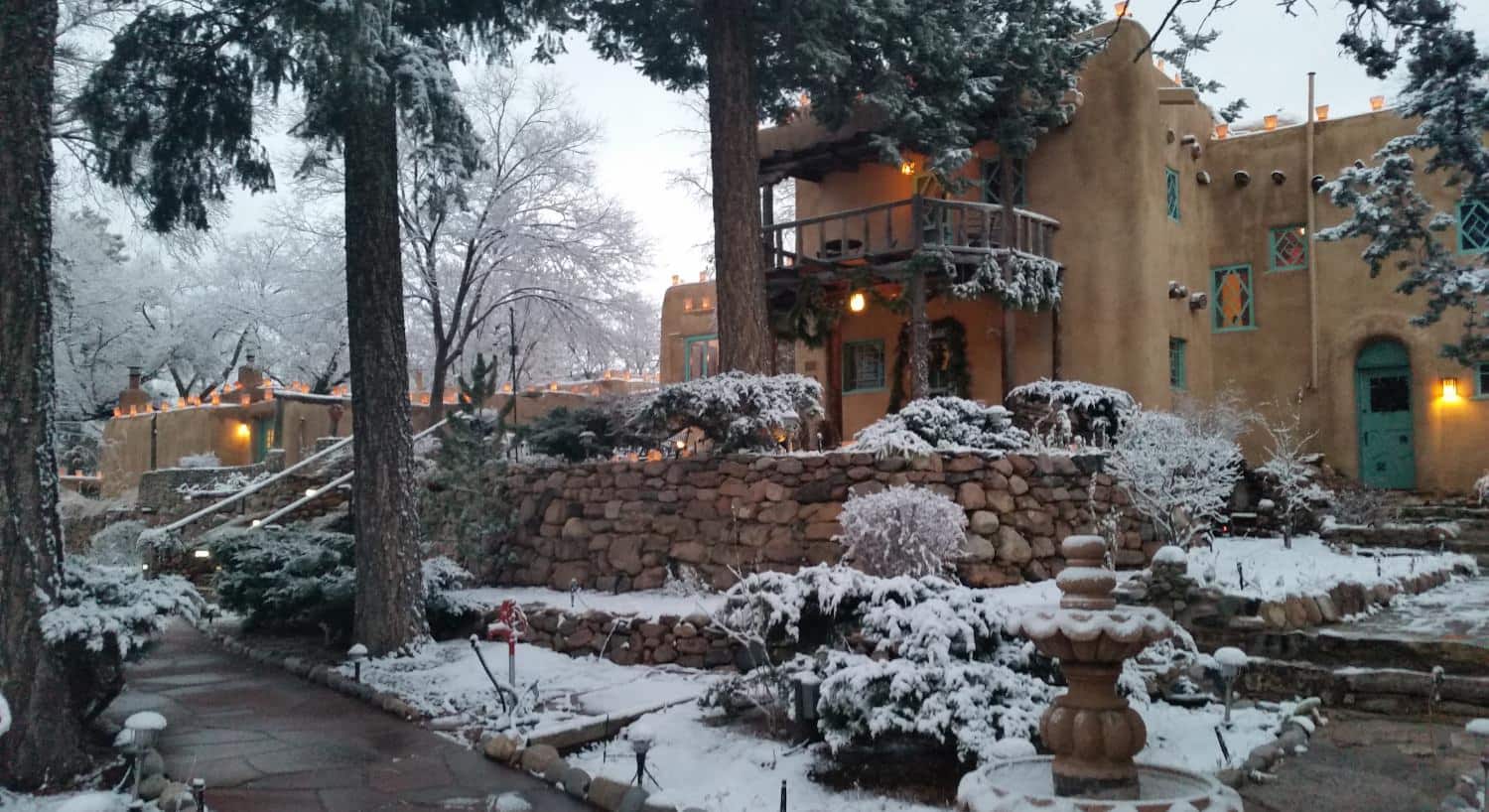 Exterior view of the property surrounded by large green trees with snow coming down, covering vegetation