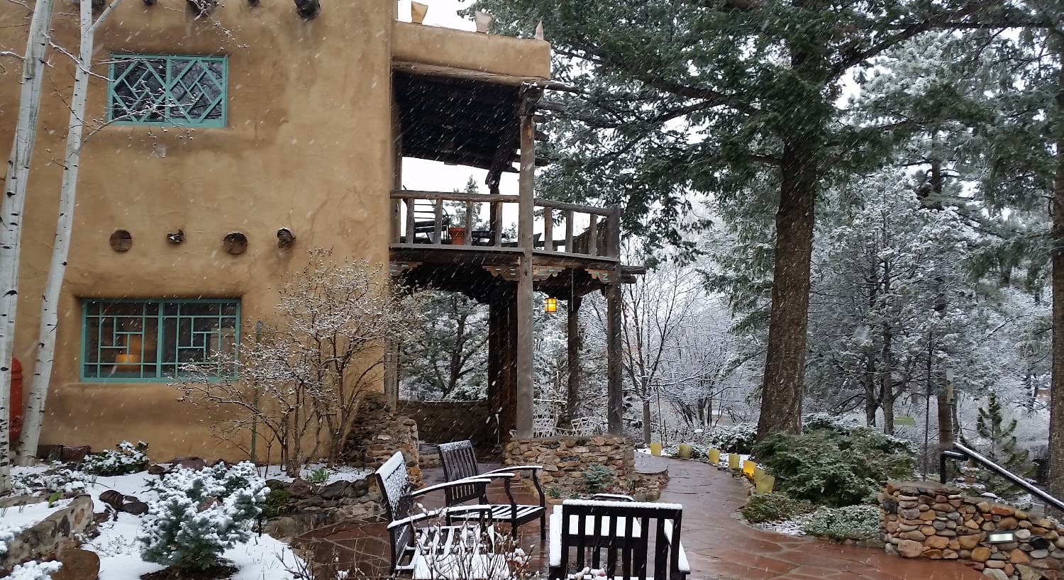 Exterior view of the property surrounded by large green trees with snow coming down, covering patio furniture and vegetation