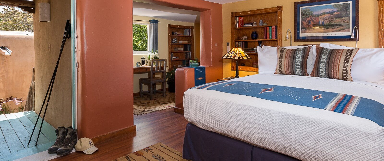 Queen sized bed with nightstands, desk and sitting room in distance, guest room door open revealing porch and entrance from the Inn's Courtyard.