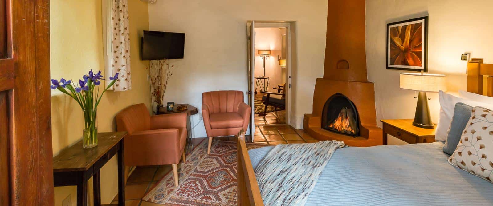 Modern-Southwestern style bedroom with fireplace, bed made up in blue and mid-century armchairs flanking a wall-mounted tv.