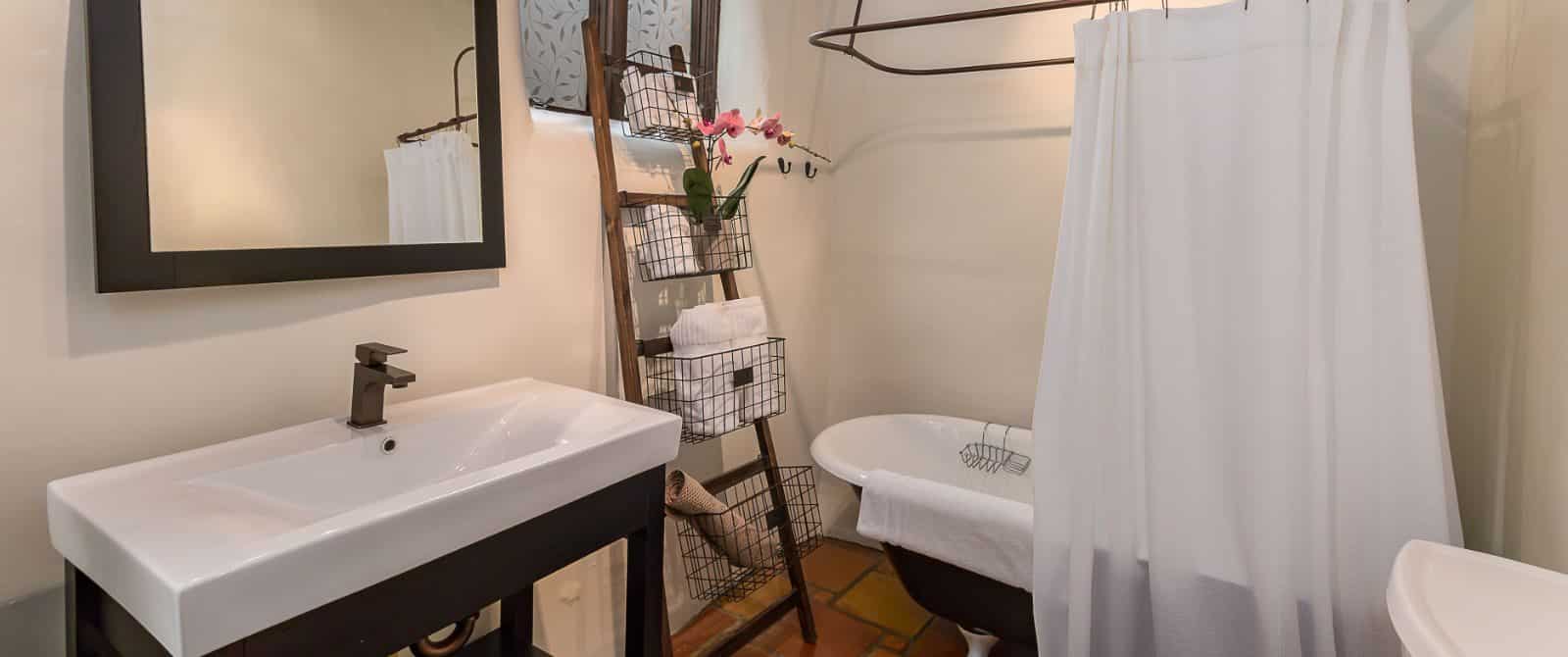 Saltillo-floor, plaster-wall bathroom with vanity in dark wood, footed tub and ladder towel rack.