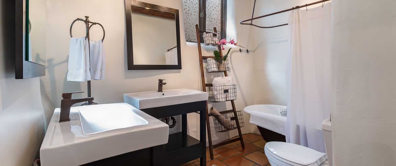 Saltillo-floor bathroom with two vanities in dark wood, footed tub and ladder towel rack.