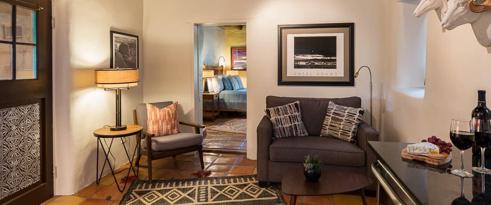 Love seat, side chair, coffee and end table, reading lamps, wine and cheese on wet bar in sitting room of two room suite. Sleeping room with queen sized bed visible through open doorway.