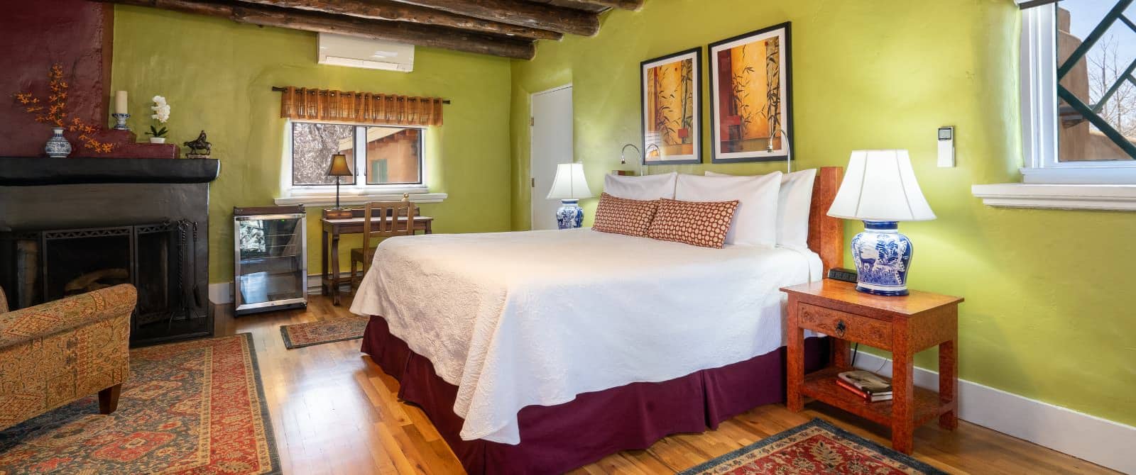 Bedroom with walls painted green, fireplace with iron grate, bed made up in white and maroon, wooden desk and chair.