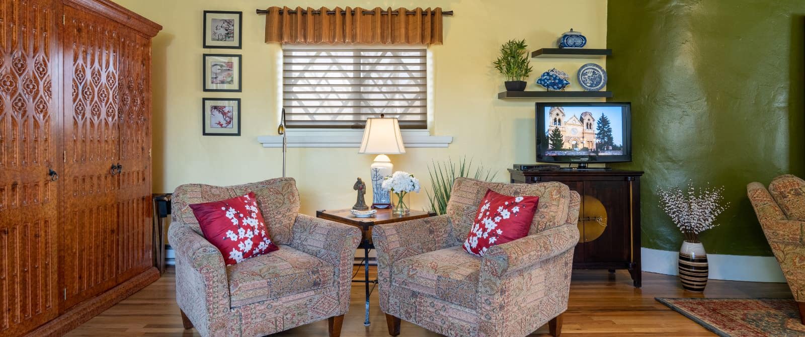 Two armchairs around wooden table in sitting area with tv, huge armoire and wooden floors.