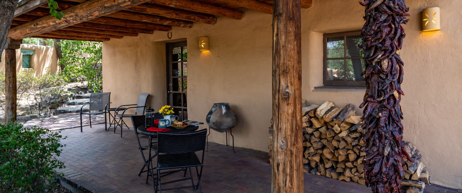 Patio under wooden overhand of adobe building with seating and stack of wood.