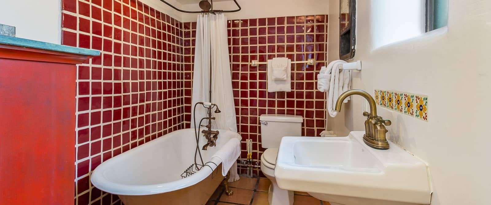 Footed tub, sink and stool in bathroom with deep-red tiles on walls and Saltillo tile floor.