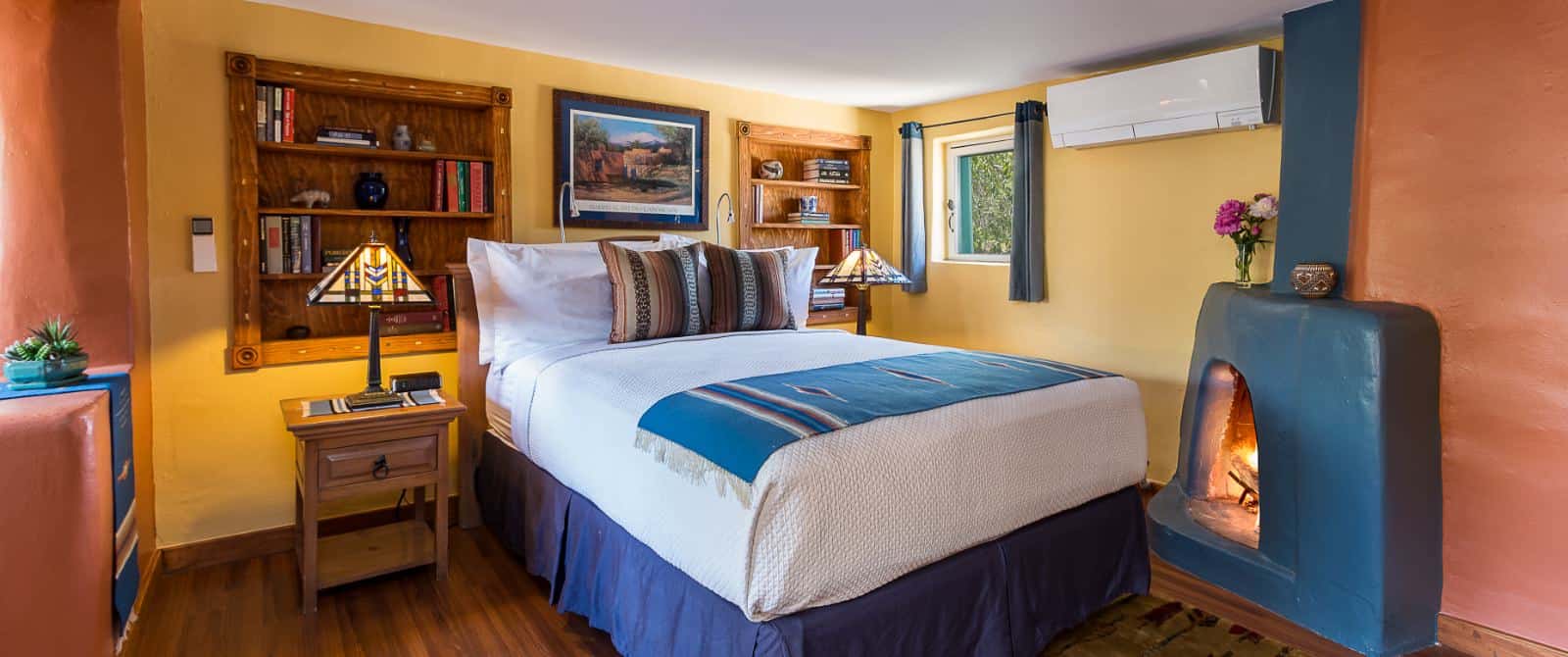 Bedroom with blue pueblo-style fireplace, bed made up in white and blue, nightstand and built-in wooden bookcases.