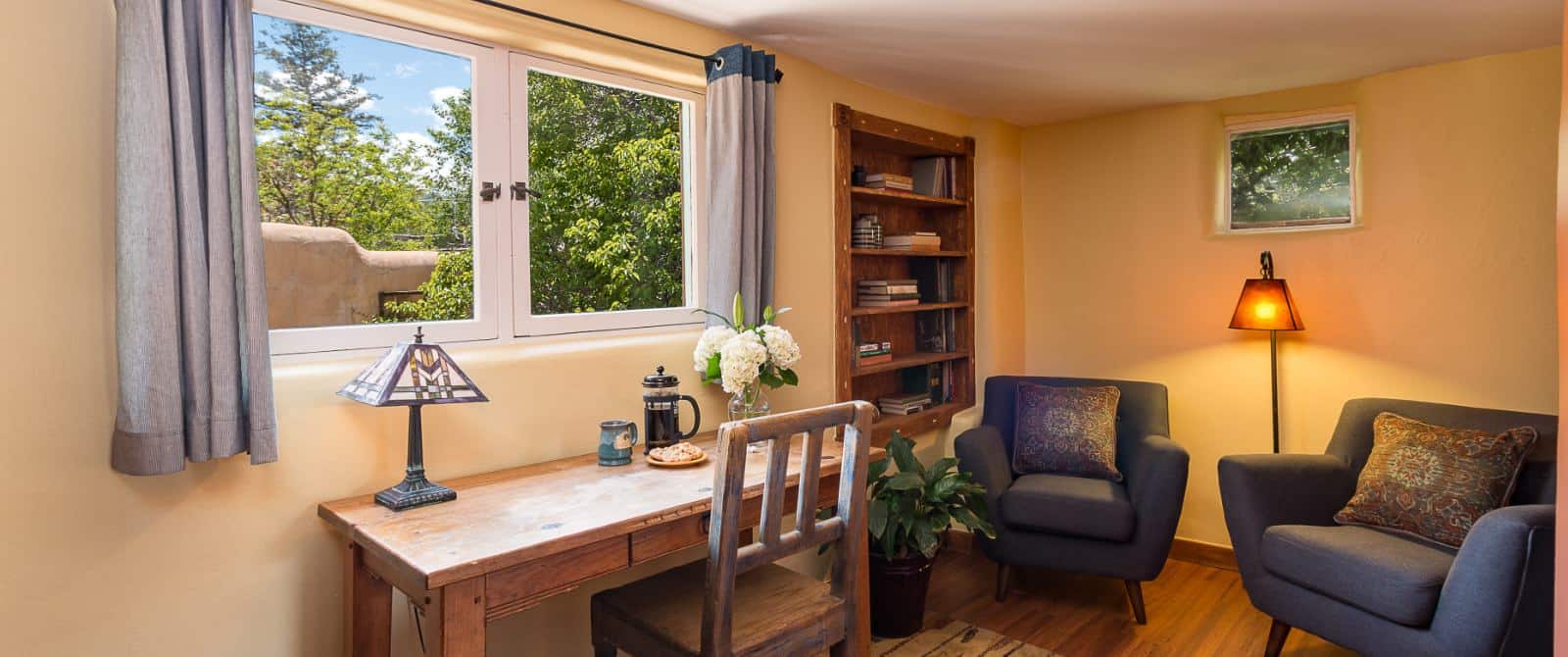 Two blue armchairs arranged for conversation next to a wooden desk under a window looking out at blue sky and green trees.