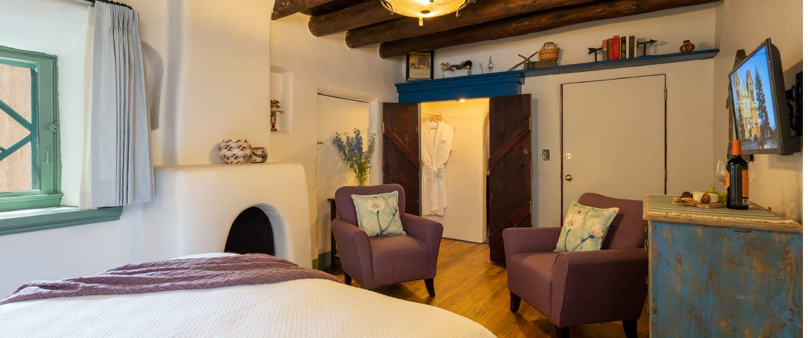 Southwest-plastered room with wooden closet doors, wood floor and wood beans in ceiling, fireplace and two mauve armchairs.