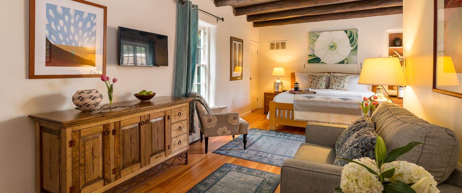 Tastefully decorated bedroom with wooden ceiling beams, plank flooring and a king bed with a sitting area including a sofa and tv.