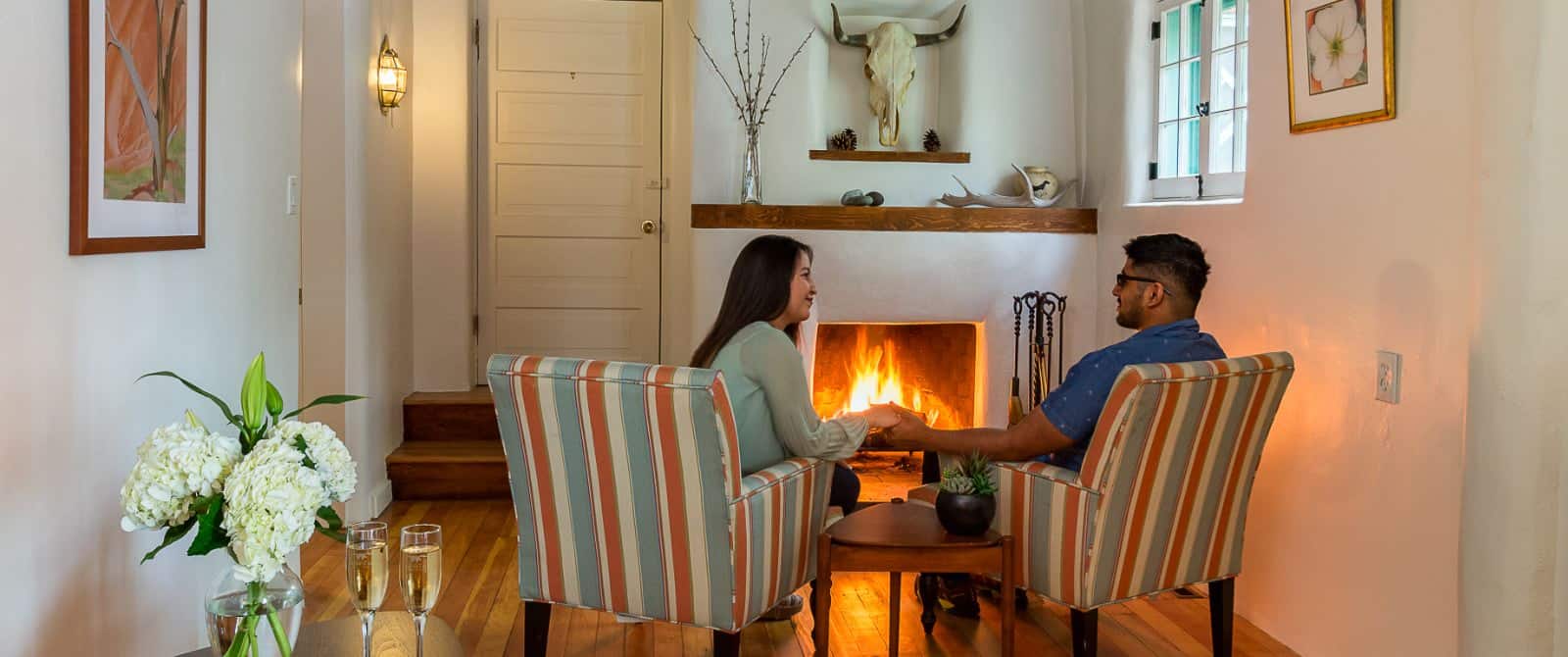 Couple sitting in two striped armchairs, holding hands over a mid-century end table in front of a southwestern-style plastered fireplace.