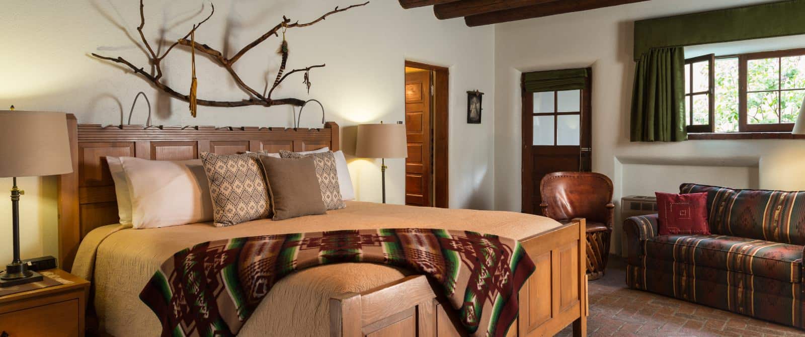 Pueblo-style bedroom with brick floor, bed made up in tan, sofa in southwest fabric, and unique wooden branch art piece.