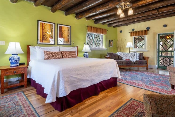 Guestroom with wooden floors, blue and red rugs, wooden furniture and bed made up in white and maroon.