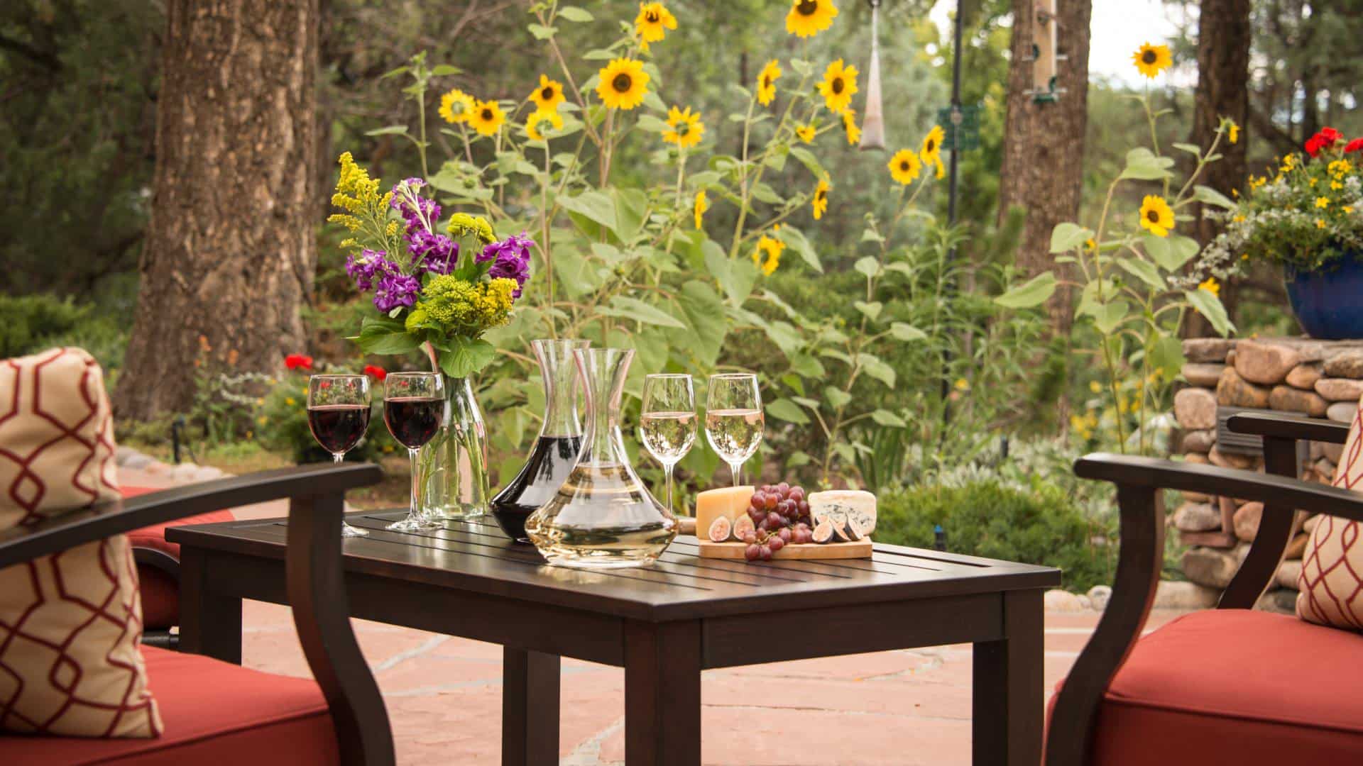 Dark wooden coffee table on stone patio with white and red wine decanters, glasses of poured white and red wine, and wooden tray with cheese and grapes