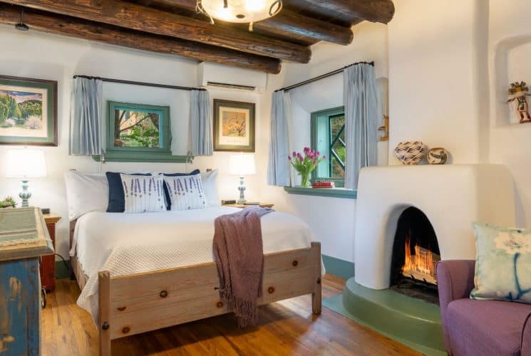 Cozy bedroom with southwest-style furniture, pueblo-style fireplace, bed made up in white and mauve armchairs.