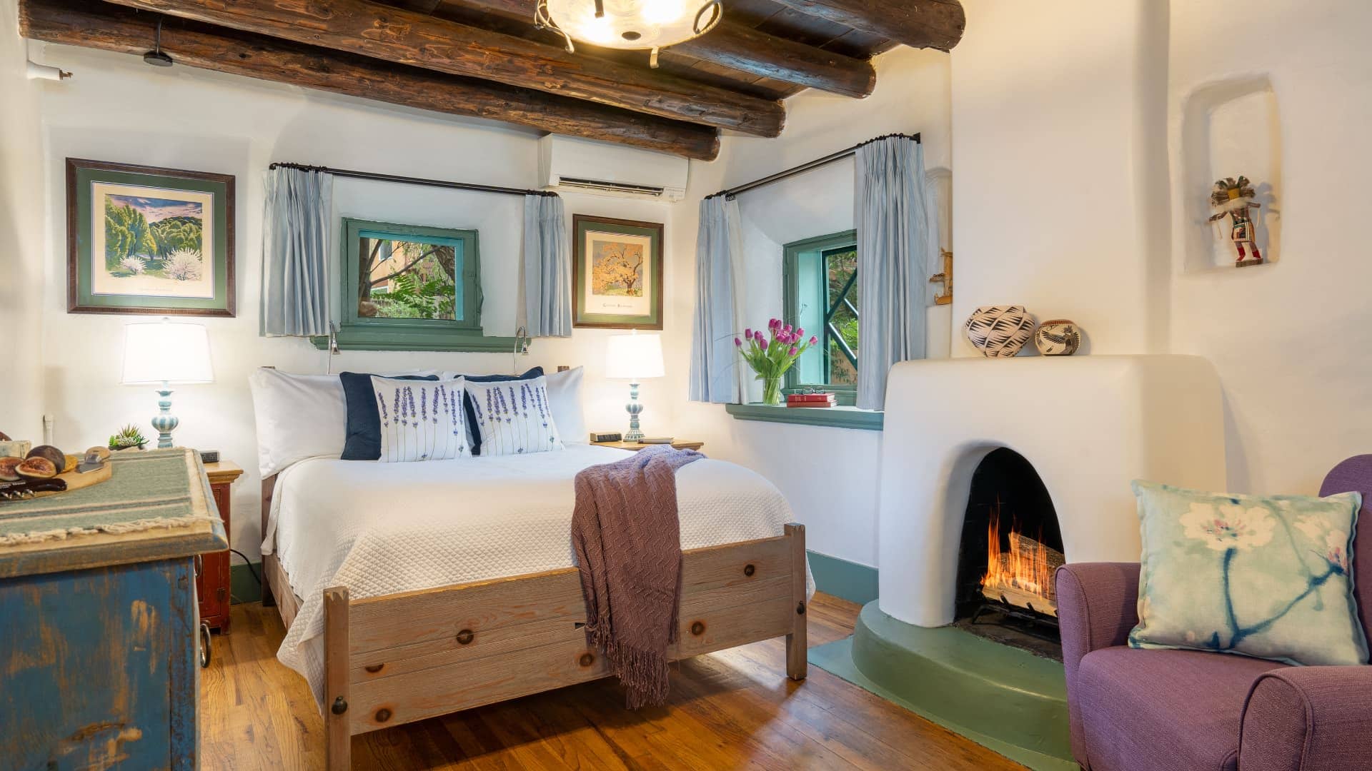 Cozy bedroom with southwest-style furniture, pueblo-style fireplace, bed made up in white and mauve armchairs.