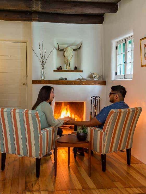 Couple sitting in two striped armchairs, holding hands over a mid-century end table in front of a southwestern-style plastered fireplace.