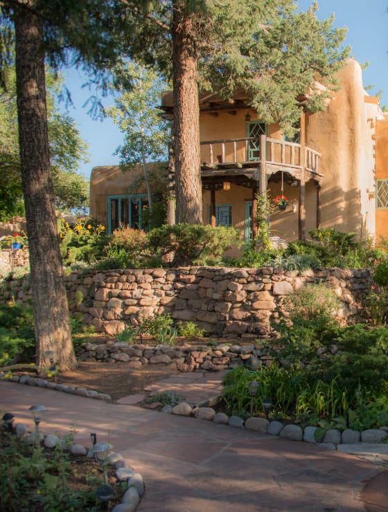 Large adobe house fronted with stone walls and walkway and large pine trees.
