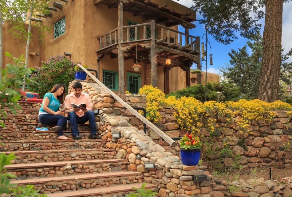 Couple reading guidebook on stone stairs outside of patio near adobe building with wooden balcony.