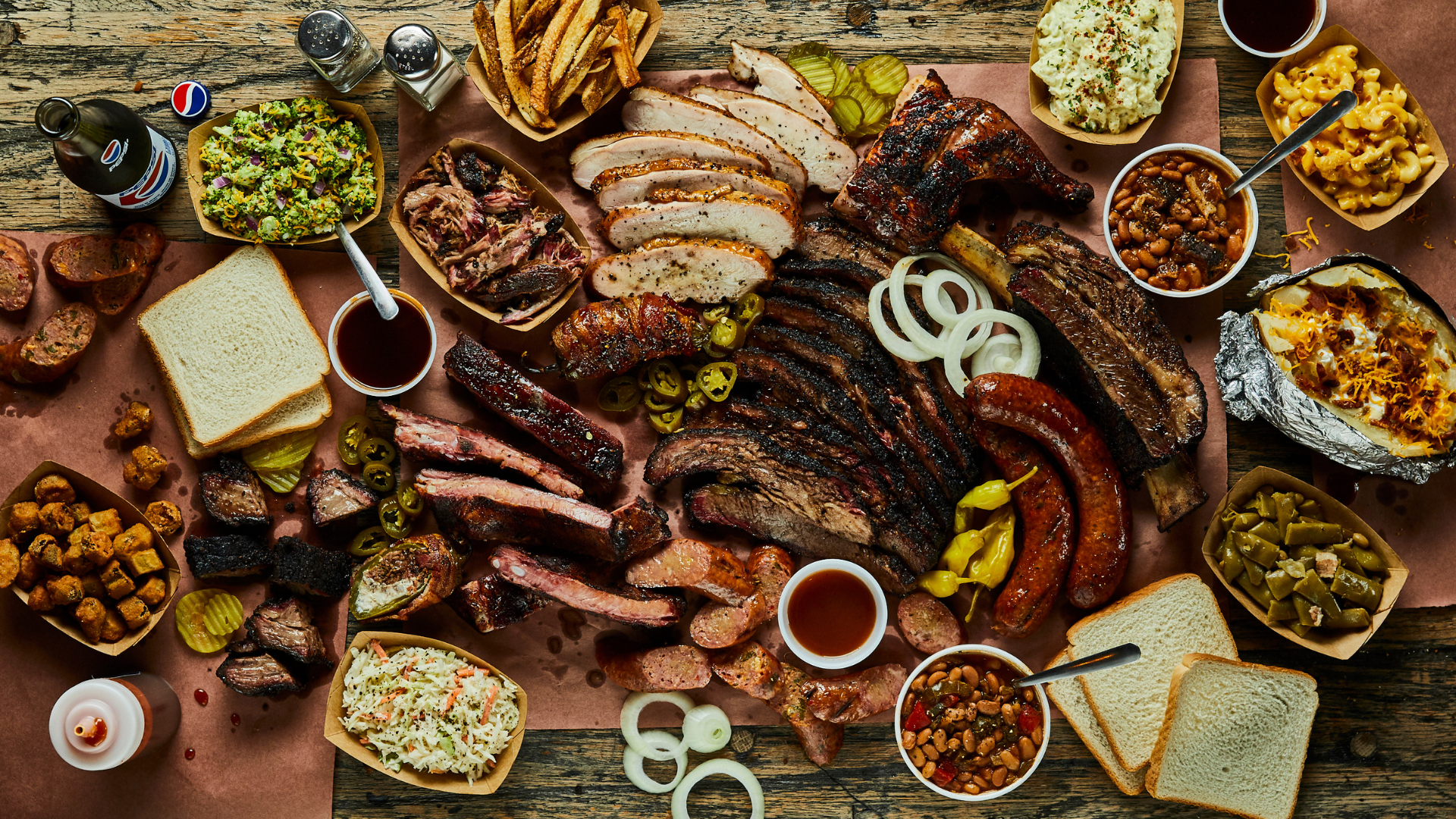 bbq spread with assorted grilled meats ribs sausages sides like coleslaw baked beans mac and cheese, and slices of white bread on a wooden table 