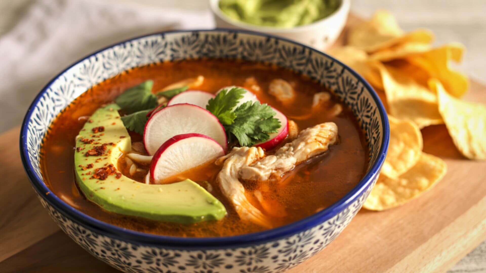 bowl of pozole topped with avocado radishes cilantro and chicken served with tortilla chips