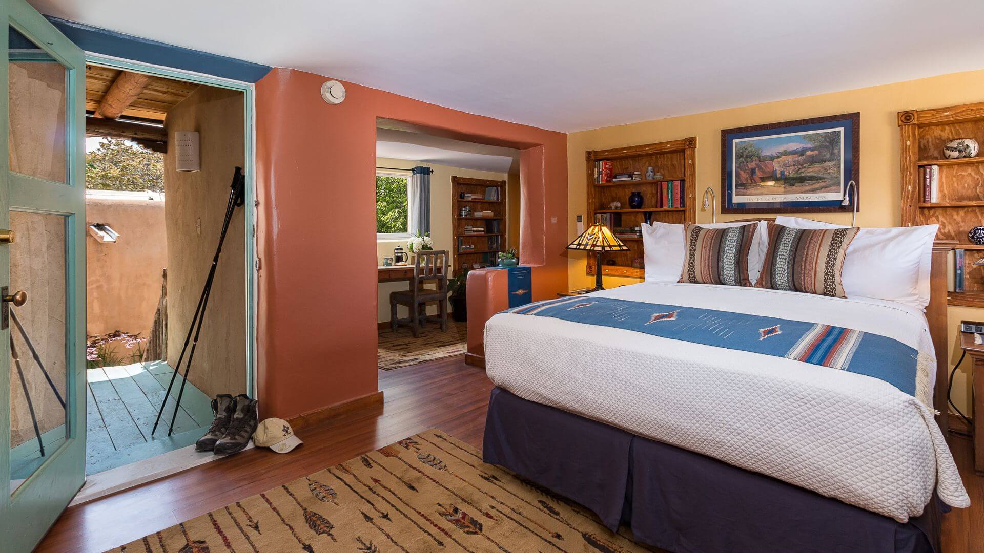 DH Lawrence guest room with a king bed, colorful textiles, wooden shelves, and a small desk area, with an open door leading to a patio and natural light filling the space