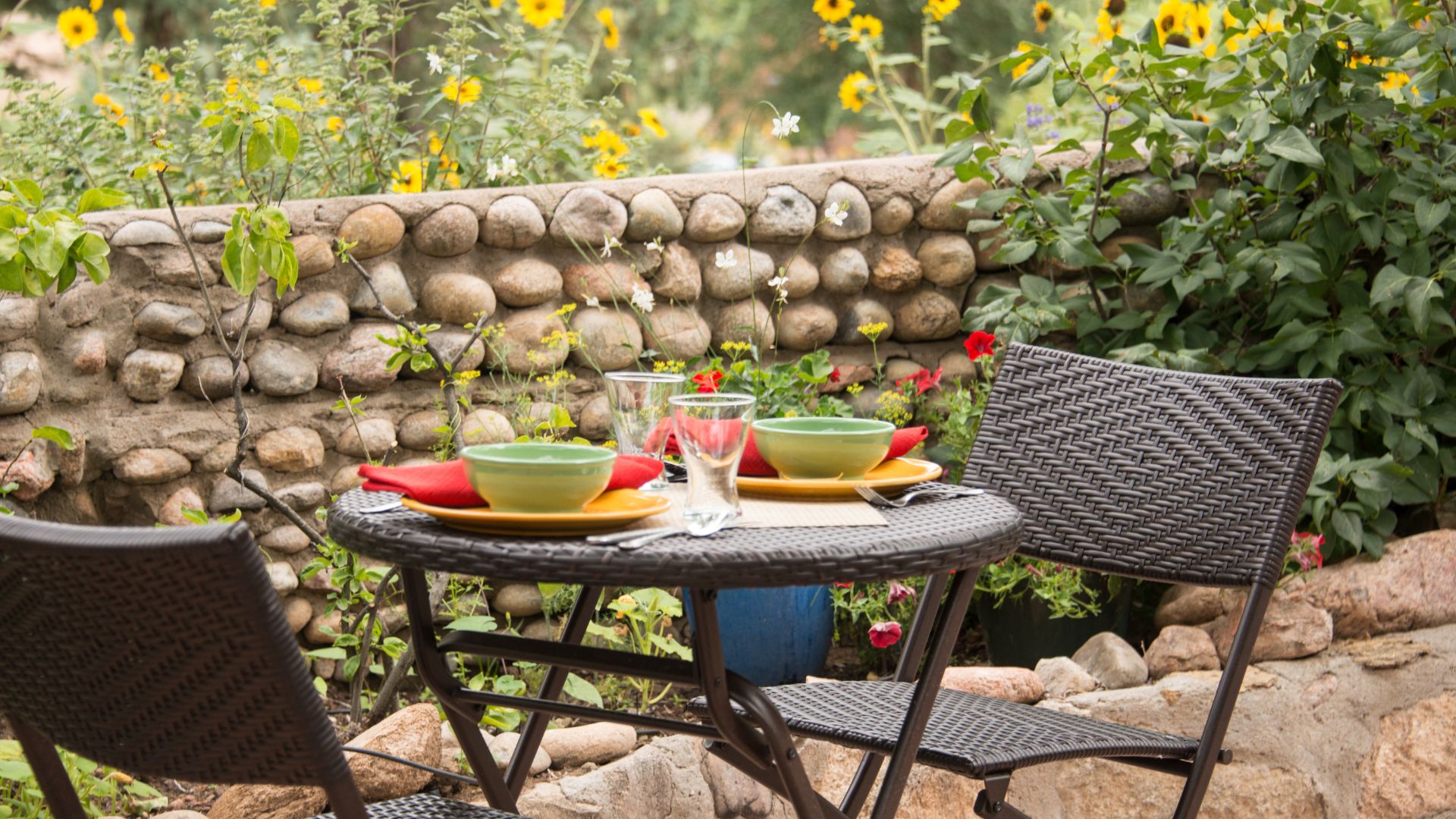Metal table for two outside on the patio surrounded by lush landscaping and set in colorful dishes ready for breakfast. 