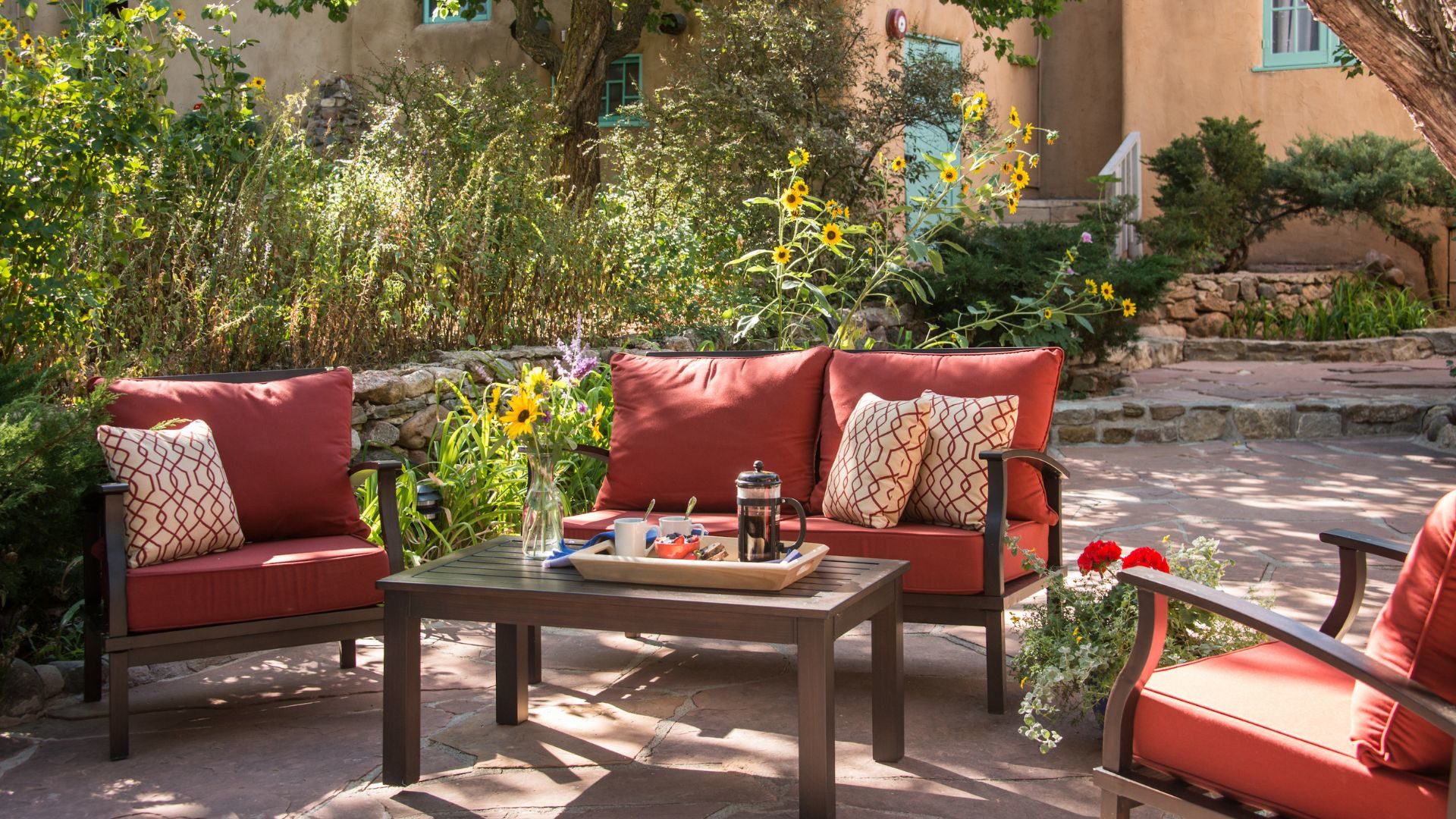 Patio furniture with rust colored cushions, printed pillows, a coffee table topped with beverages, surrounded by lush landscaping