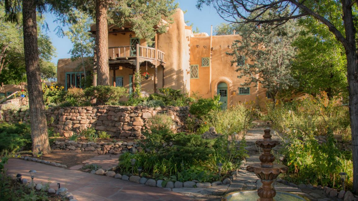 Exterior view of adobe two-story inn with portico, lush foliage and stone paths