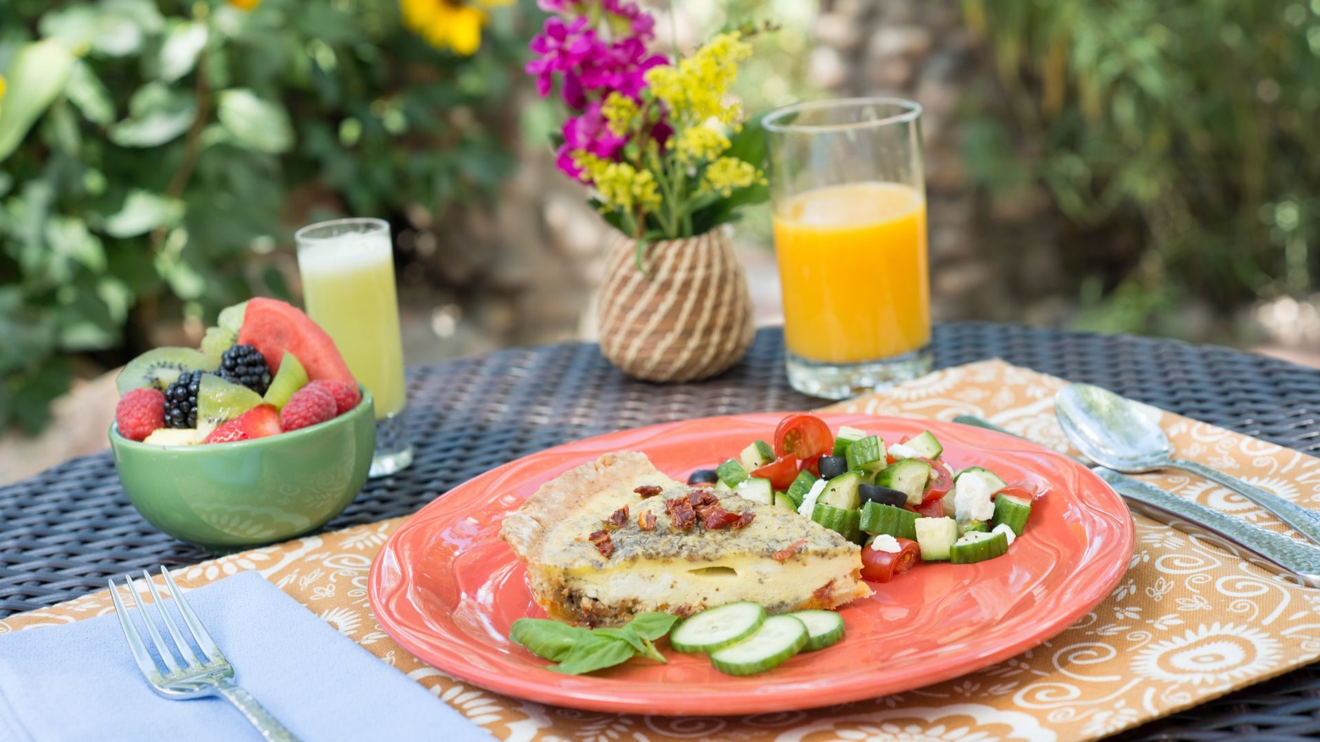 Orange stoneware plate topped with quiche and veggies, a bowl of fresh fruit and a glass of orange juice - all in a beautiful outdoor setting