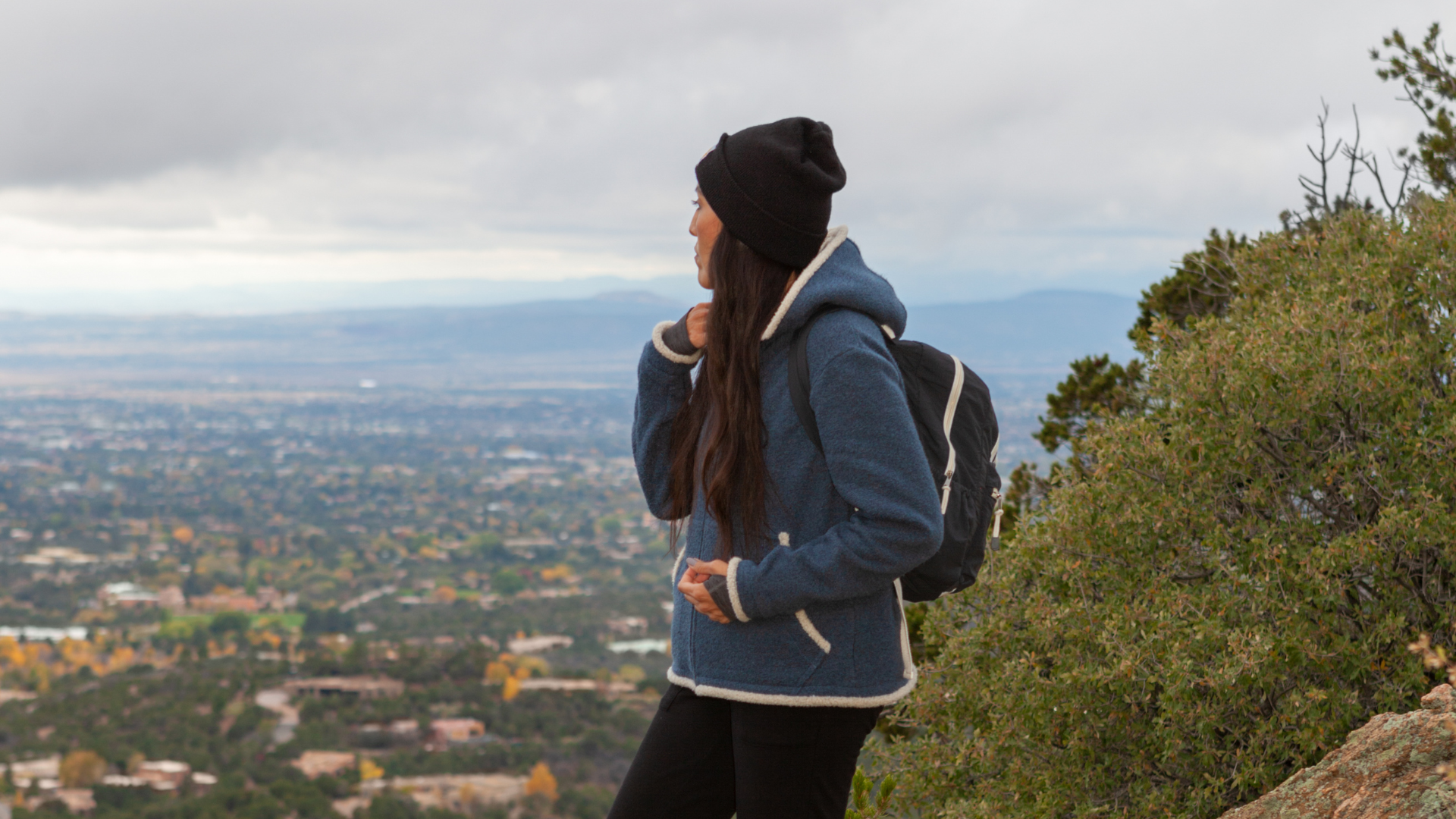 person wearing a beanie and backpack standing on a hillside overlooking a city and distant mountains
