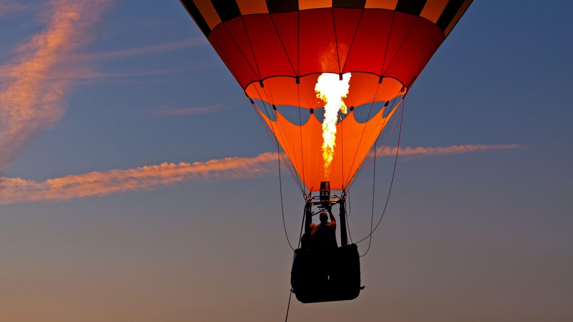 A hot air balloon glows as its burner fires against an evening sky.