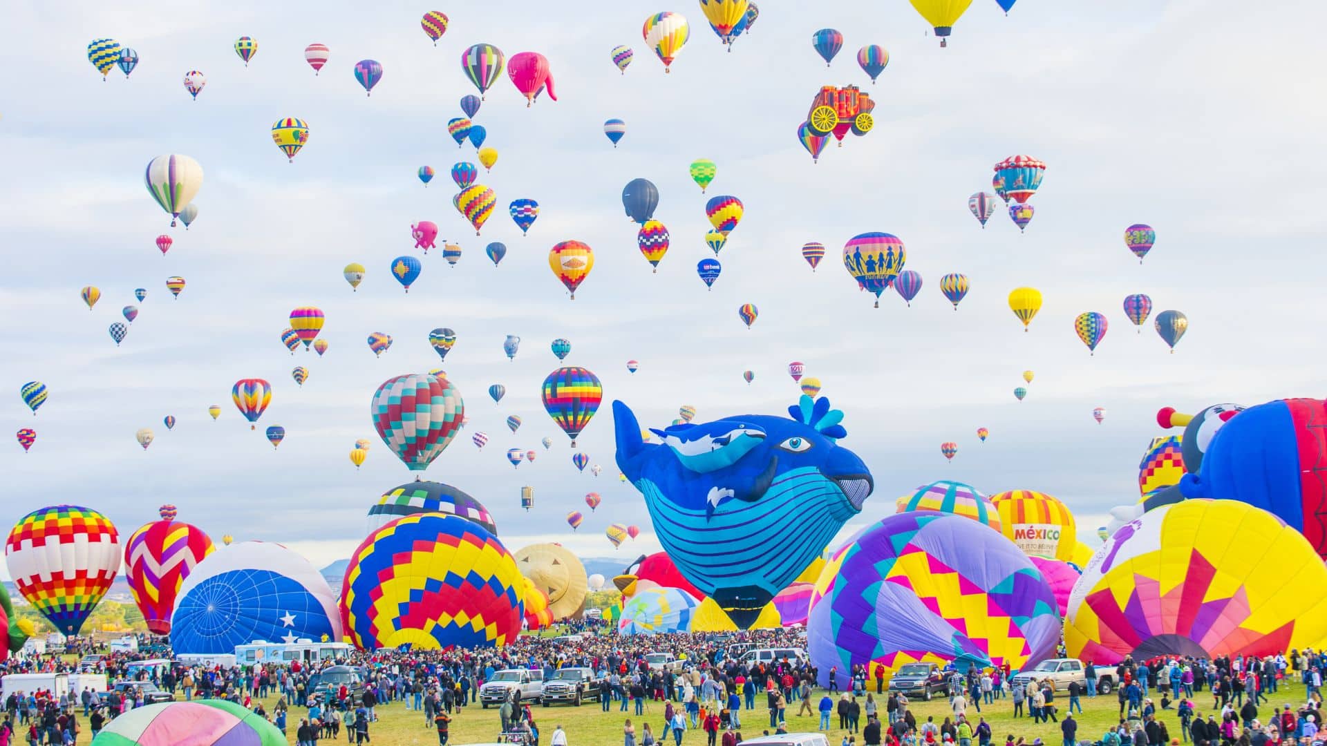 Array of brightly colored hot air balloons floating up towards blue skies with soft white clouds and crowds on the ground