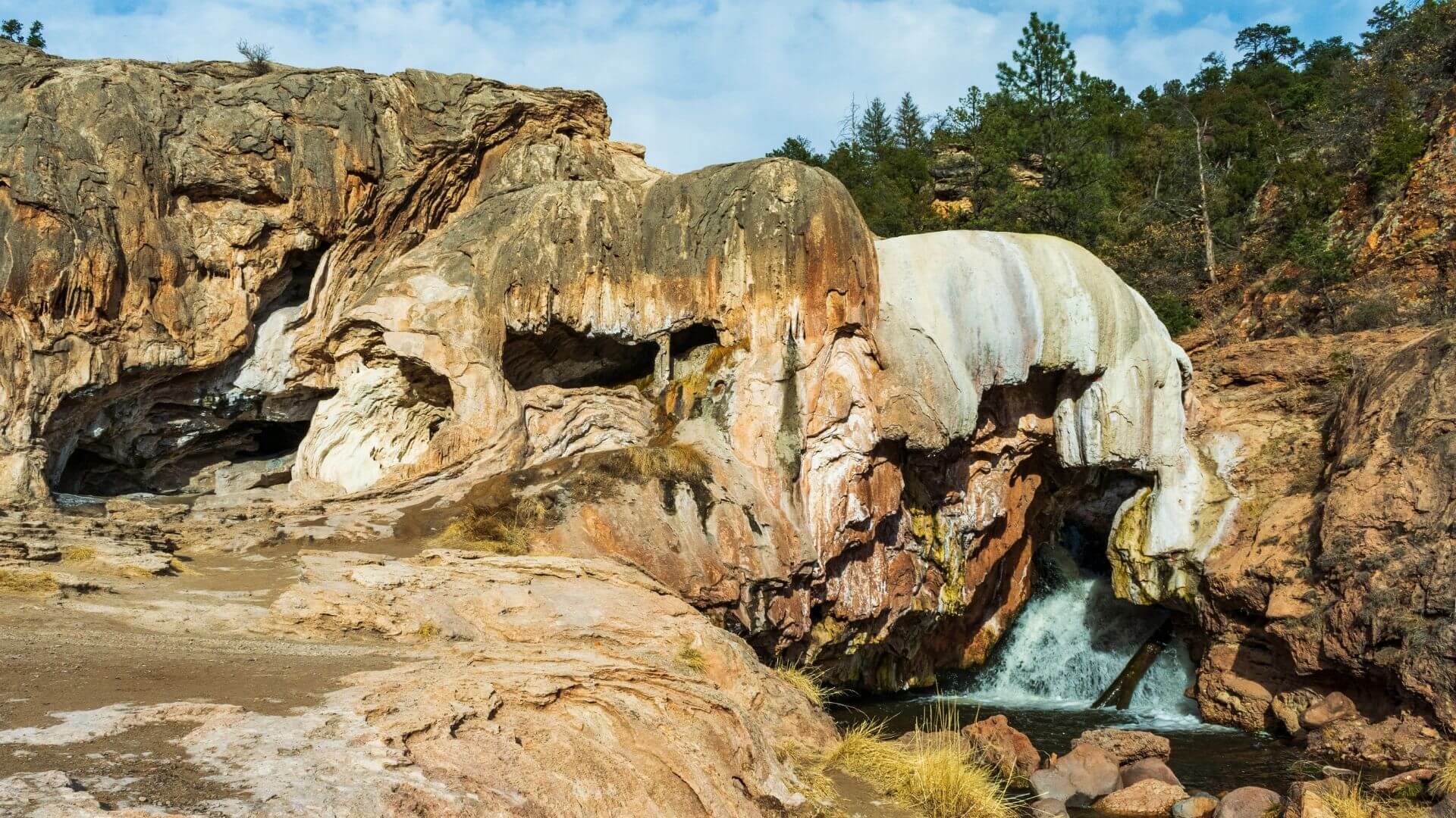 A rocky waterfall flowing through mineral formations, with rugged cliffs, pine trees, and a bright sky above